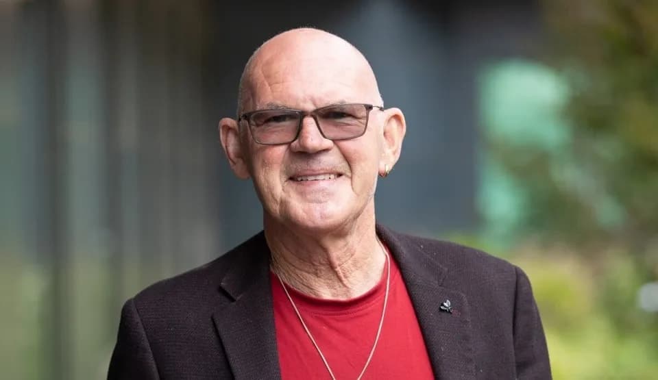 A man in tinted glasses and a red shirt smiles at the camera
