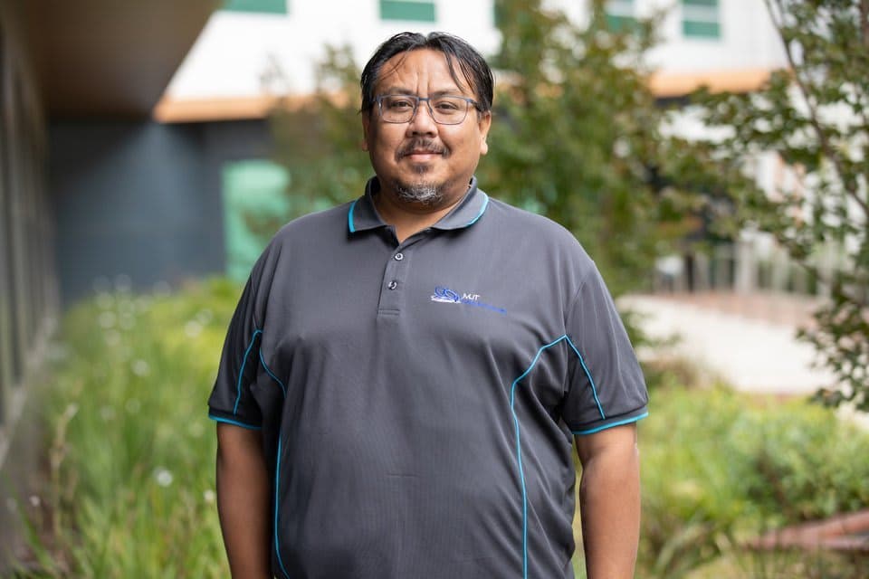 A man in a grey top standing with trees behind him