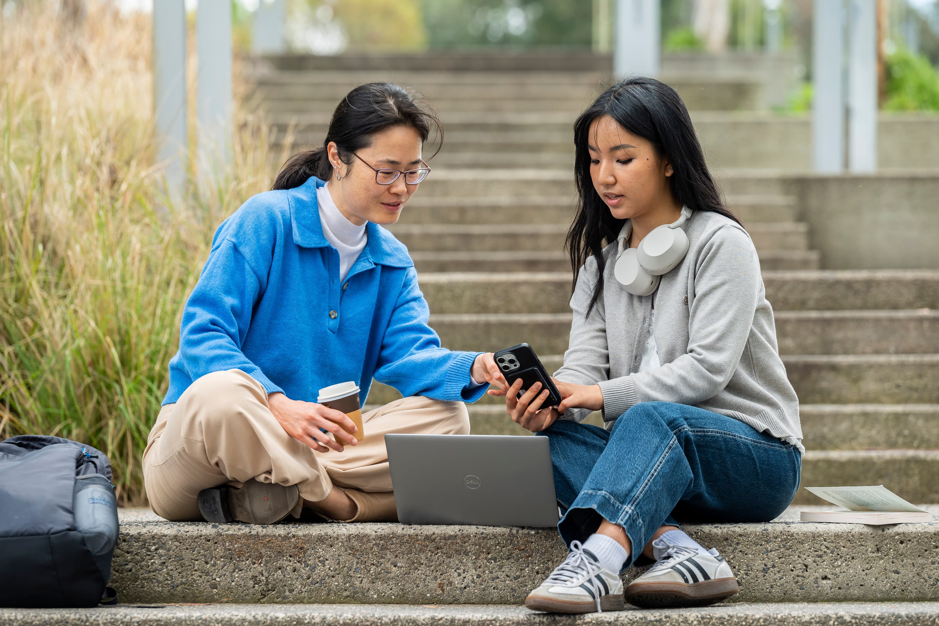Two students sit on a concrete staircase with a laptop open and look at a mobile phone that one of the students is holding.