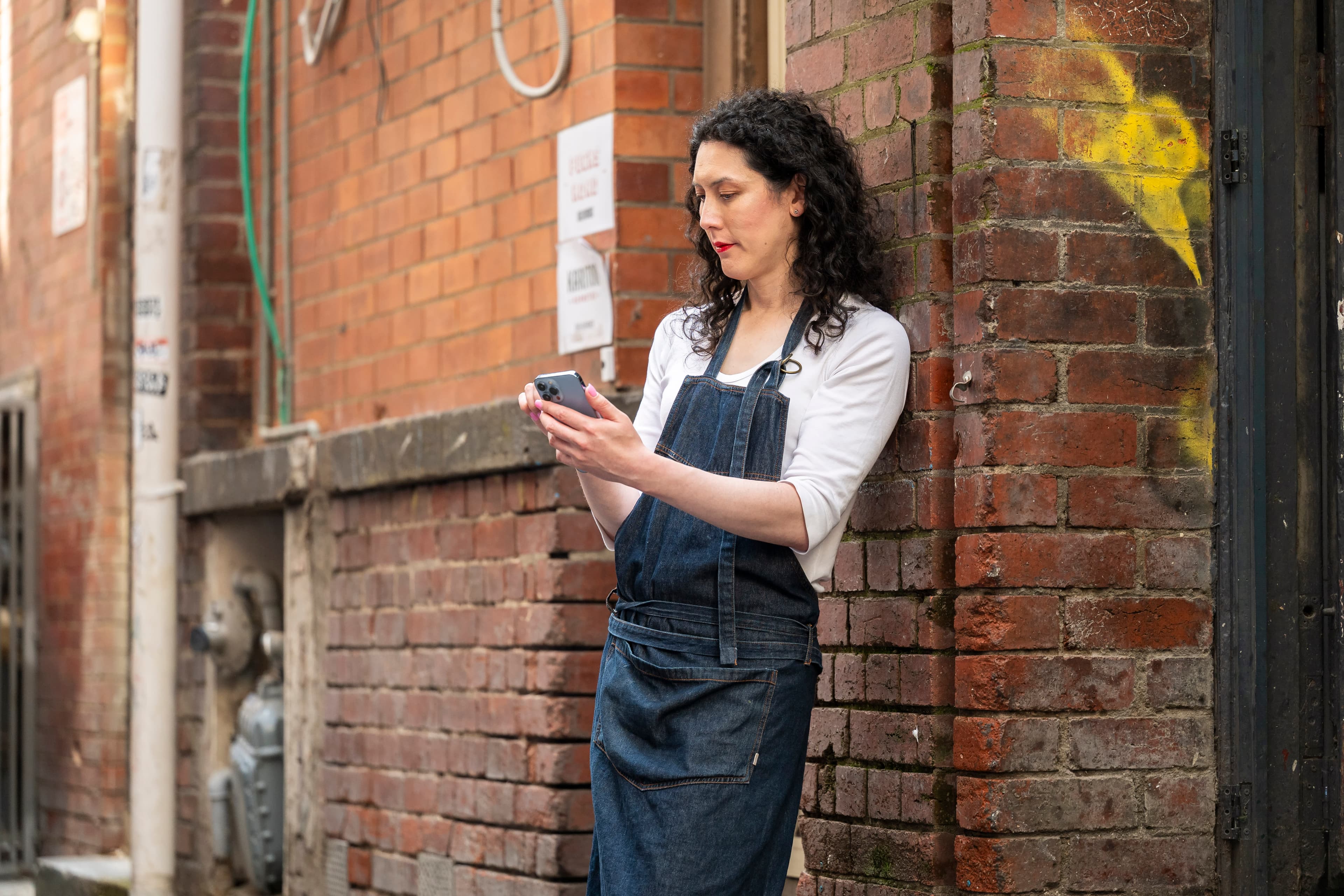 A woman with black curly hair wearing a which shirt and black apron looks at the mobile phone she is holding while leaning against a brick wall.