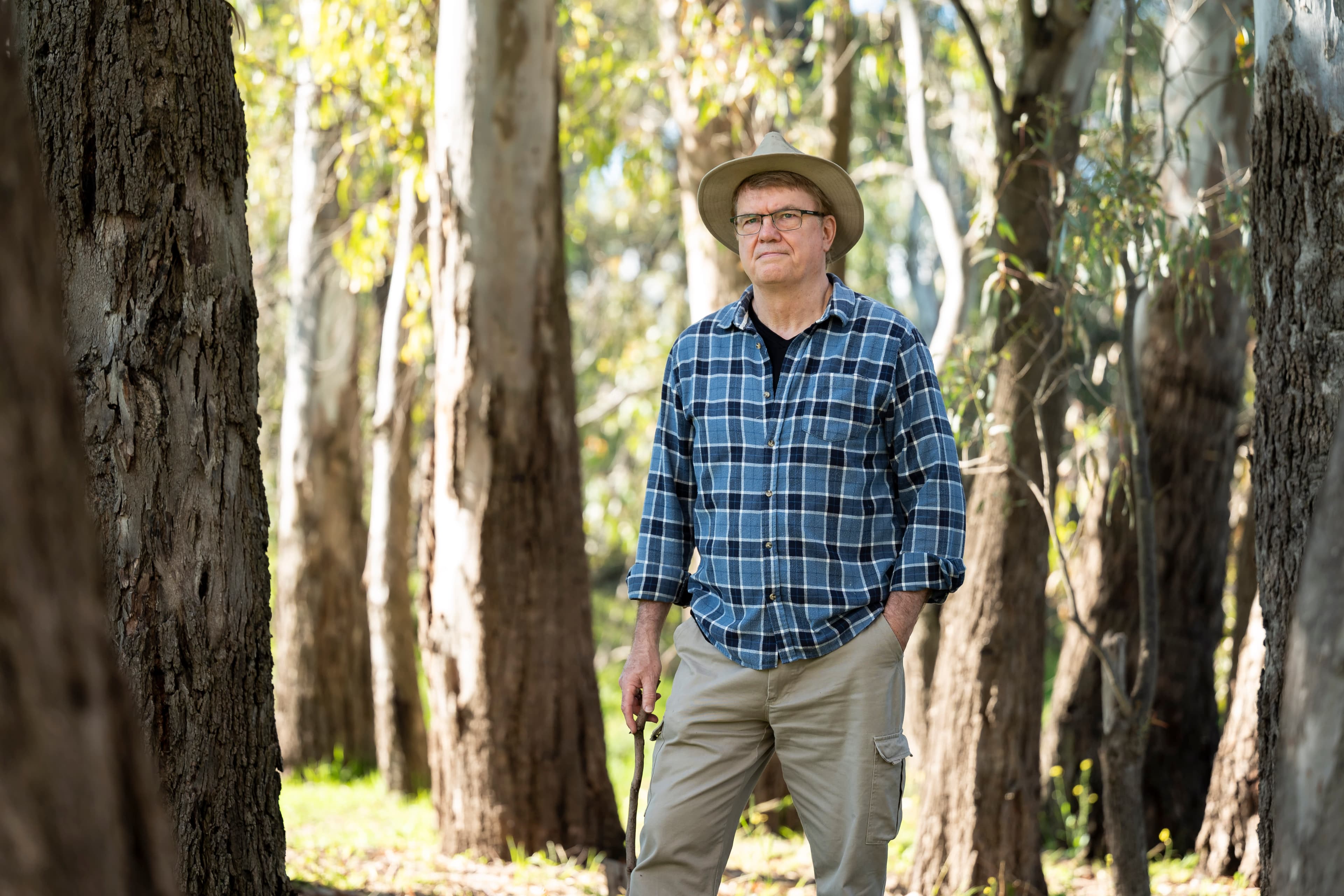 A man stands in the bush holding a stick and wearing a wide-brimmed hat, checkered shirt and tan pants.