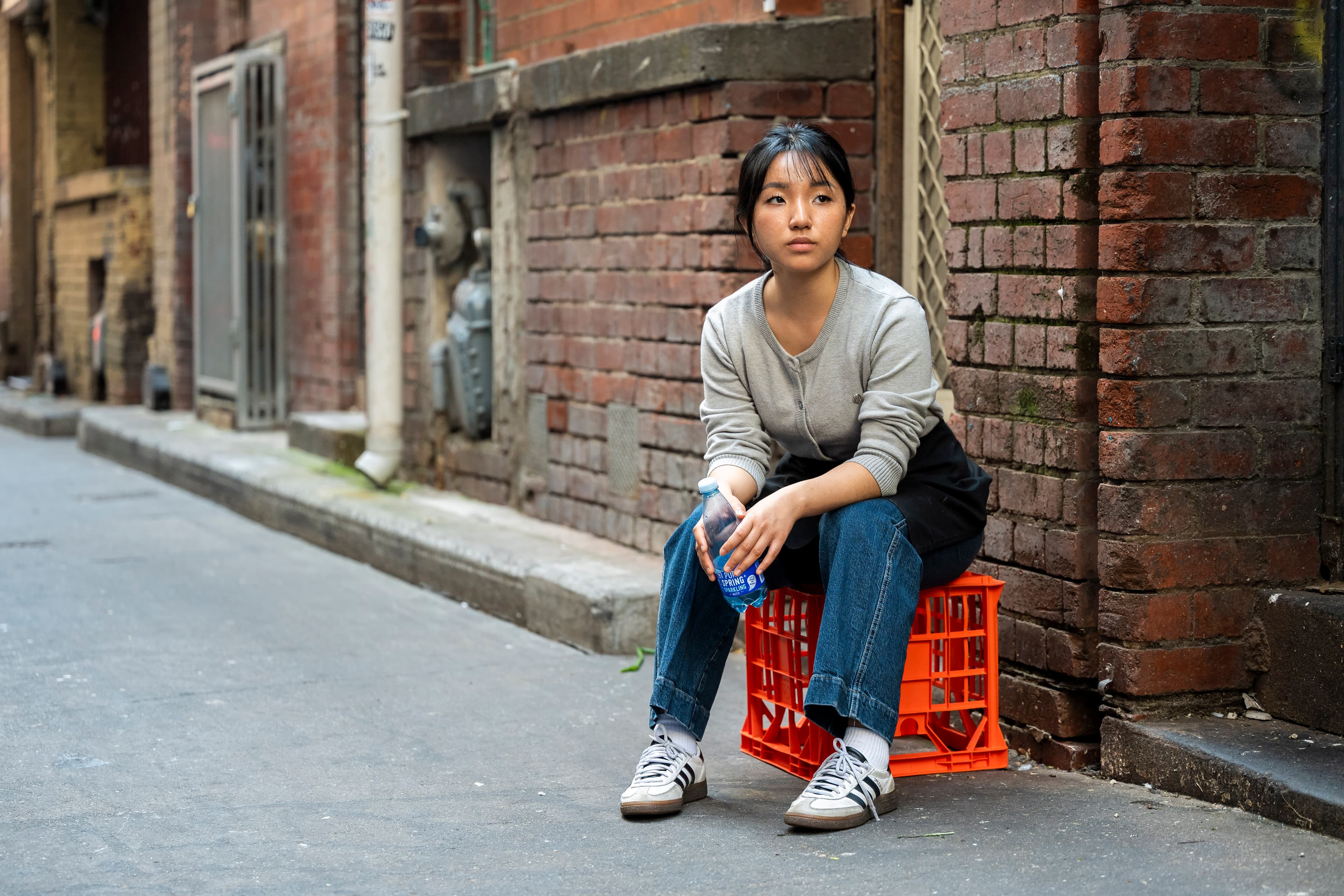 A woman wearing an apron sits on a red milk crate in a laneway looking into the distance.