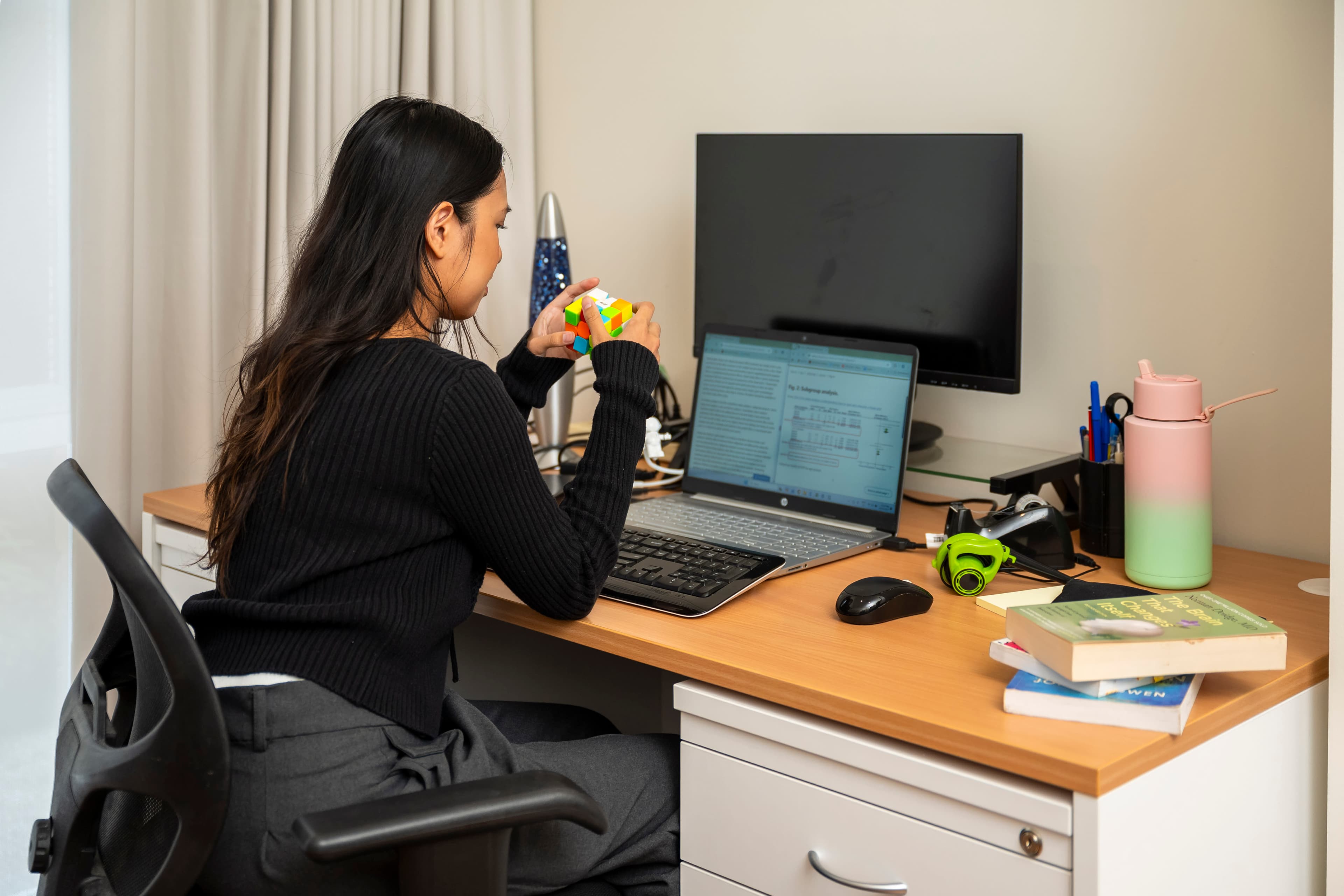 A young woman with long black hair sits at a desk facing an open laptop and holding a rubik cube.