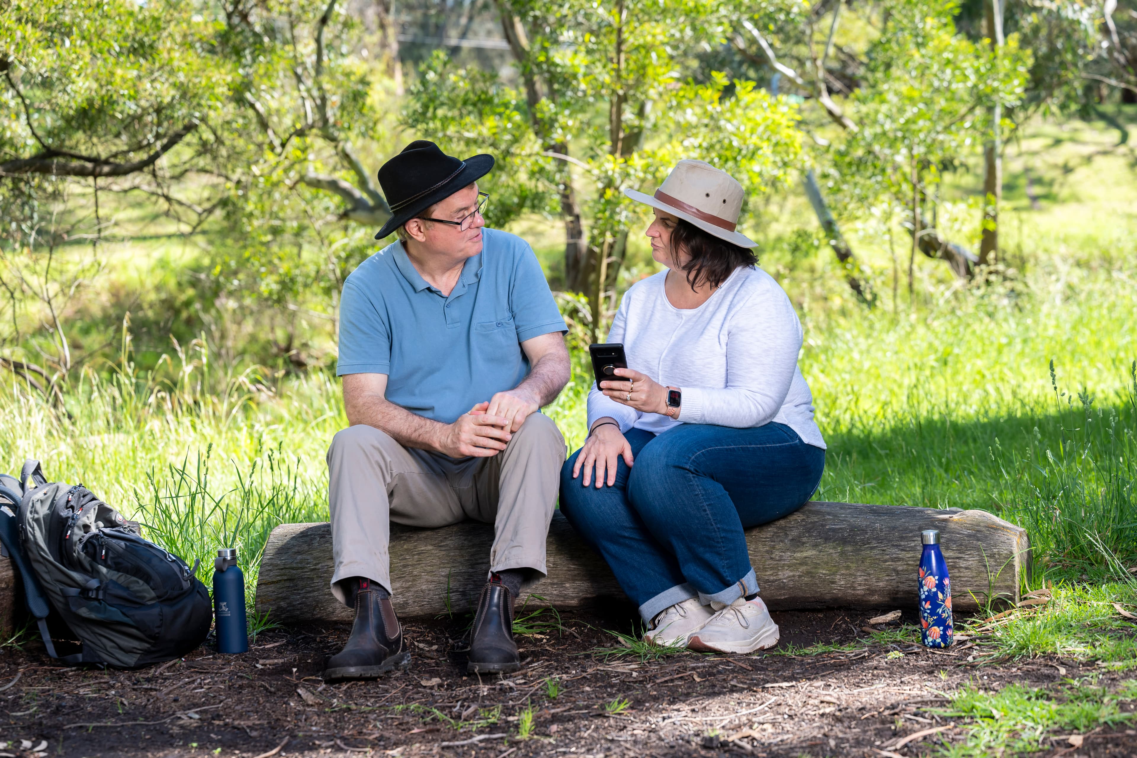 A man and a woman wearing wide-brimmed sunhats sit on a log in the bush talking to each other.