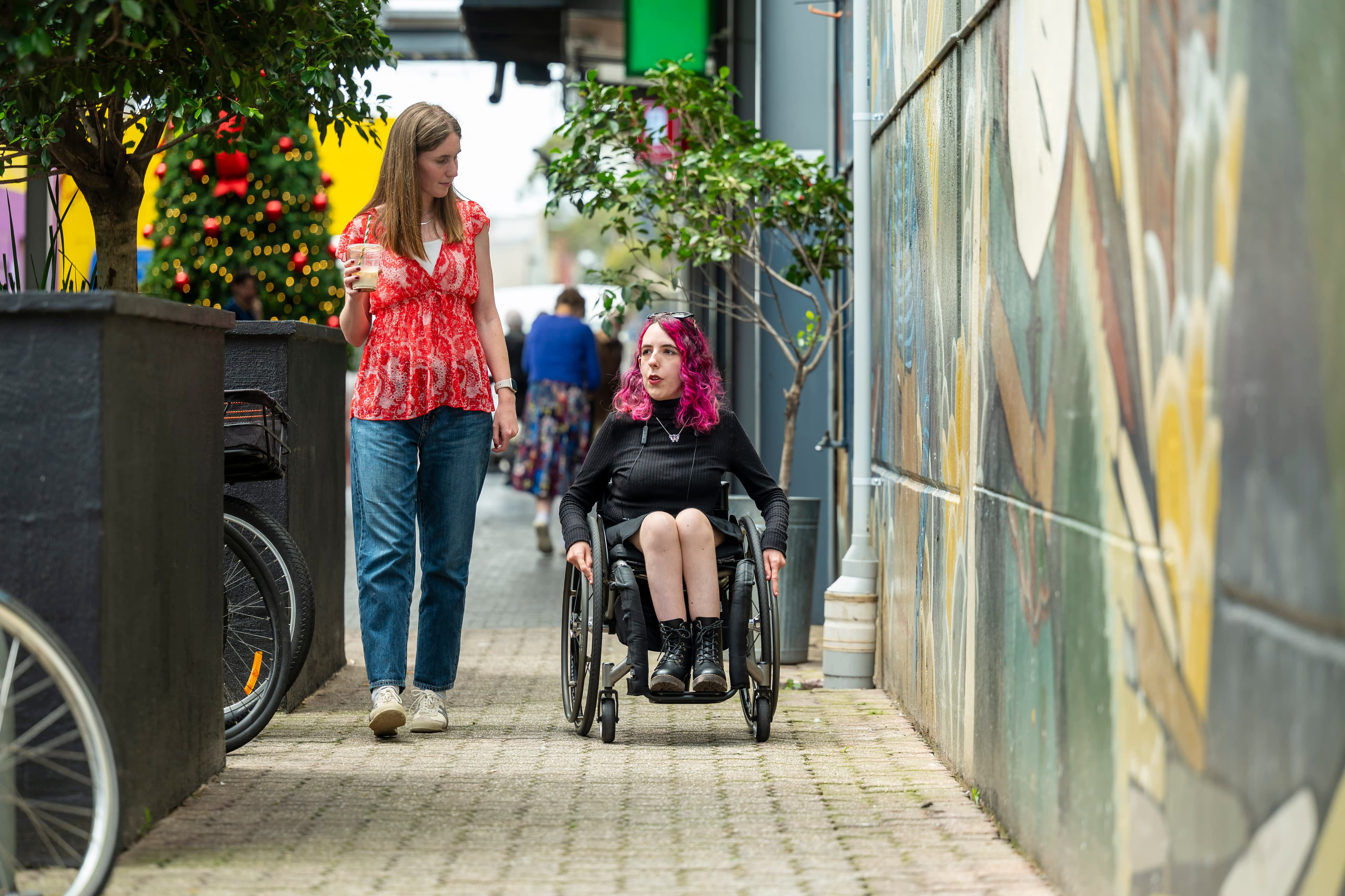 A blonde woman dressed in a red top and denim jeans walks alongside a woman with bright pink hair and lack top and skirt operating a wheelchair.