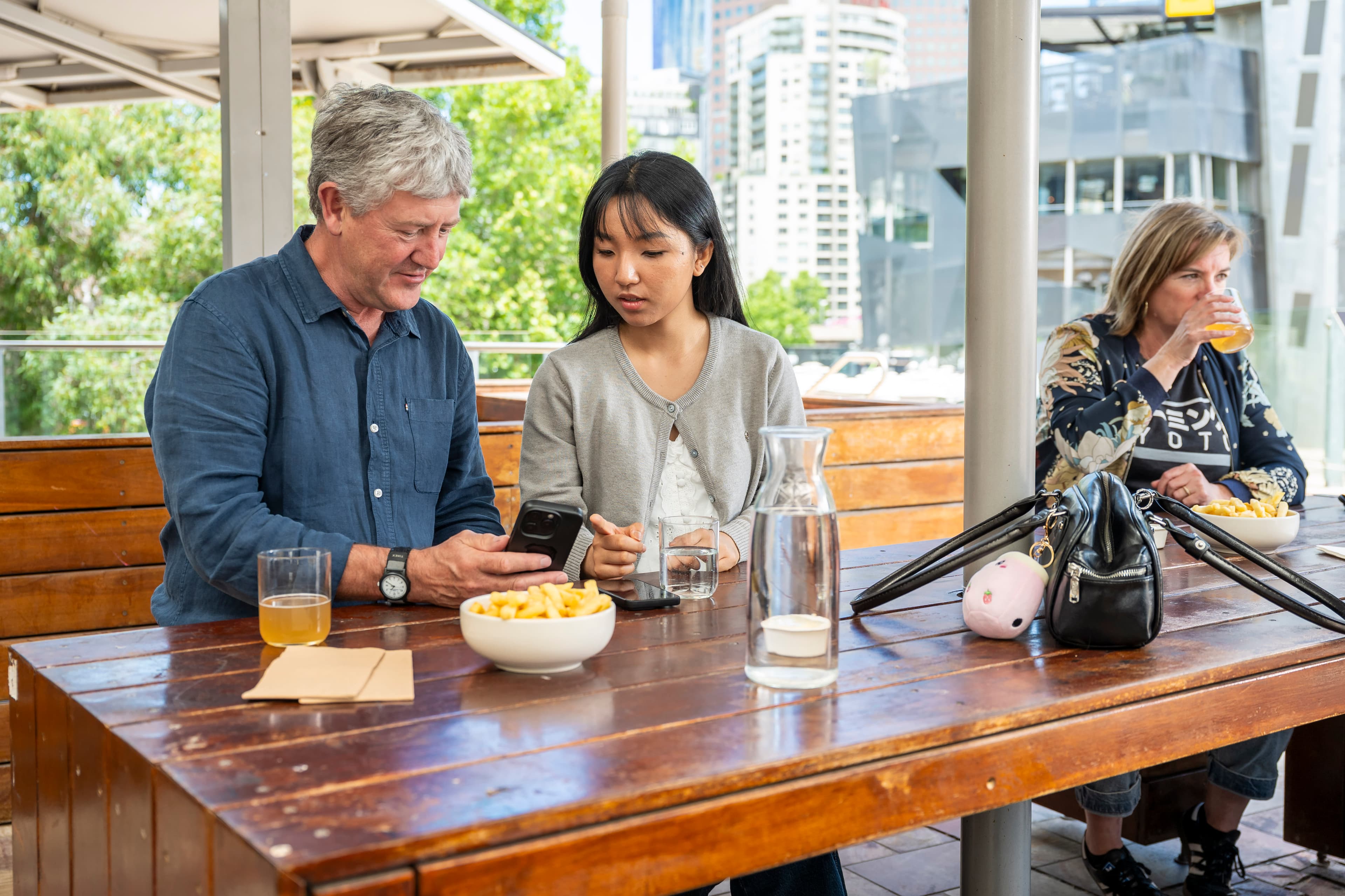 A man with grey hair and wearing a blue shirt looks at a mobile phone that he is showing to a young woman sittig next to him at a wooden table with black hair and a grey cardigan.