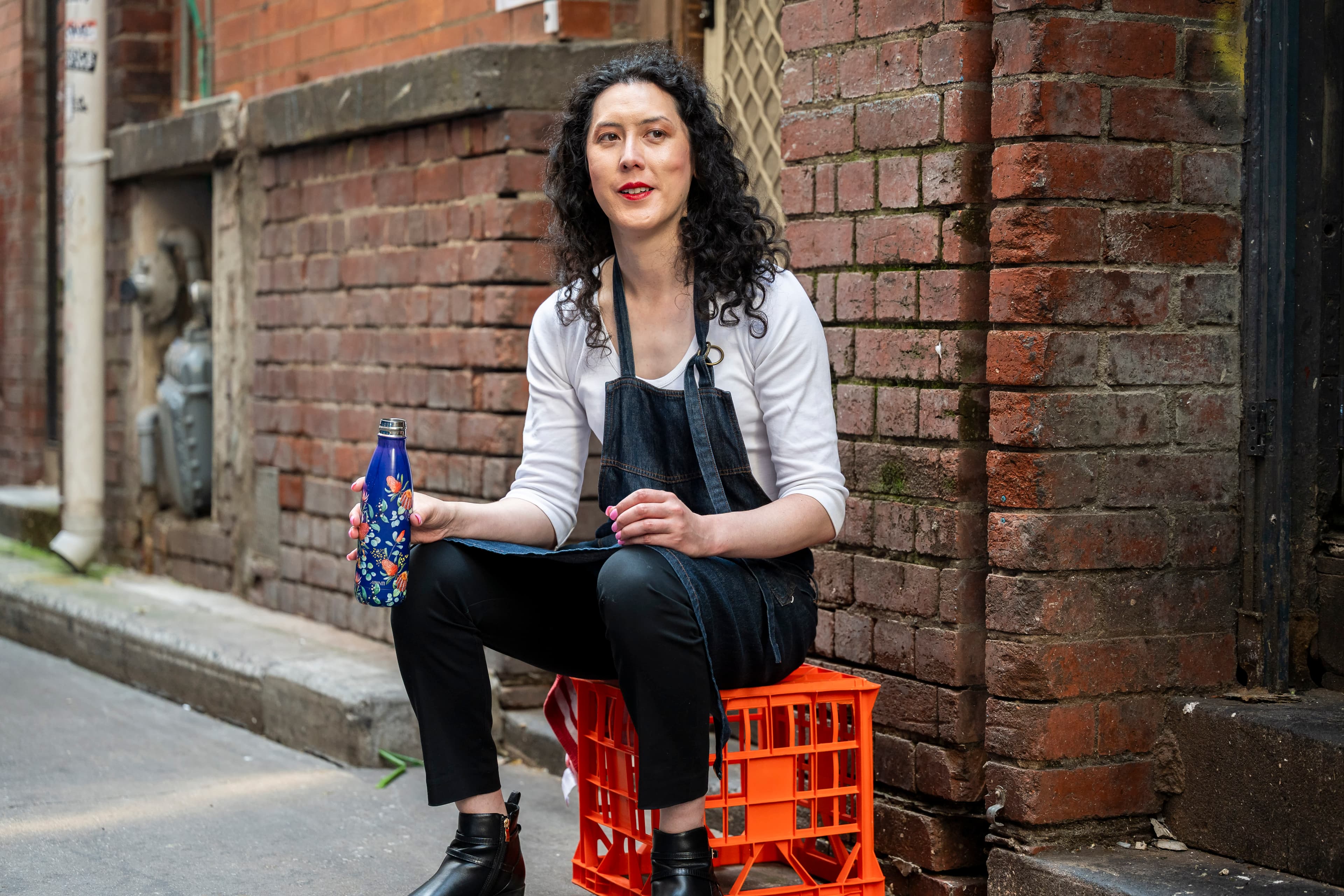 A woman wearing an apron and holding a drink bottle sits on a crate in an alley.