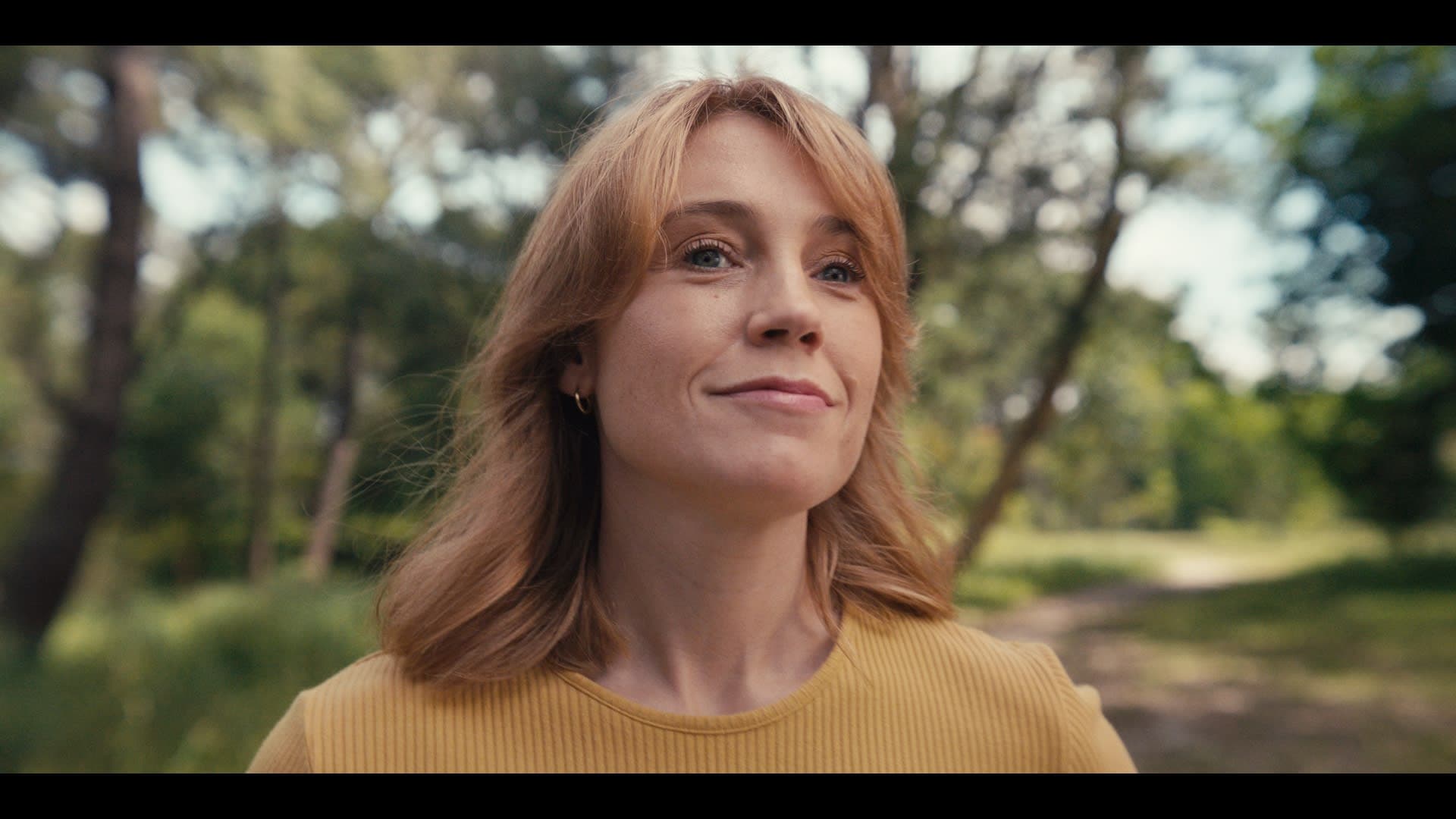 A red-haired woman in an outdoor setting smiles thoughtfully.
